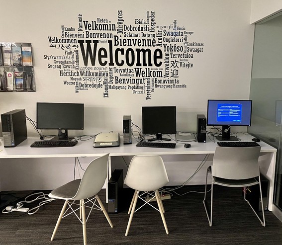 Three empty computer work stations are clustered beneath a word cloud depicting "welcome" in many different languages, but in which the English word is significantly larger than any other language.