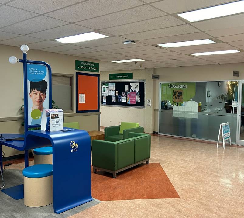 A sitting area outside of a room labeled the "global lounge." In the foreground is a prominent blue desk advertising banking services with an image of a young, winking man and the slogan "Start a conversation that matters." Another sign on the wall behind the blue desk reads Indigenous Student Services. 