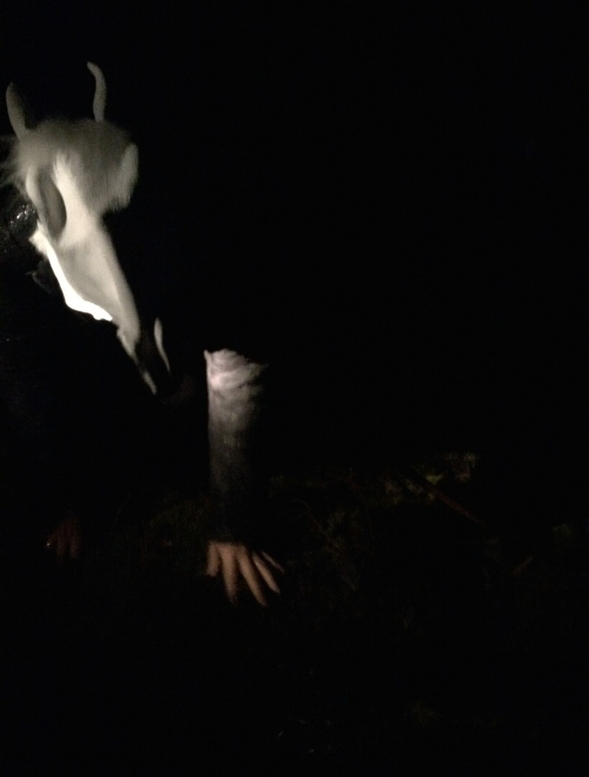 A person with a white horse mask on her head, kneeling in the dark