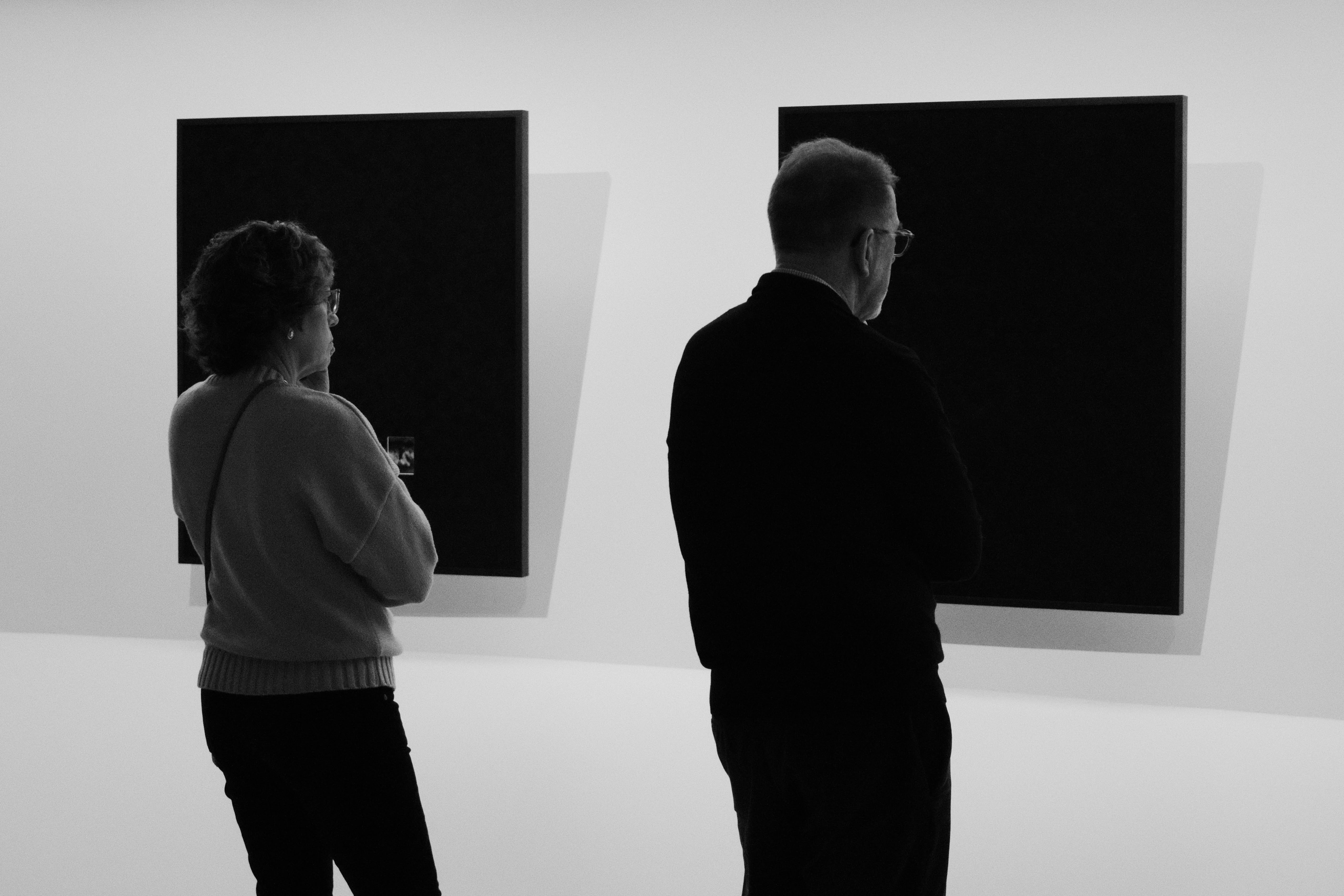 Black and white photo of two people studying two black quares/artworks on the wall