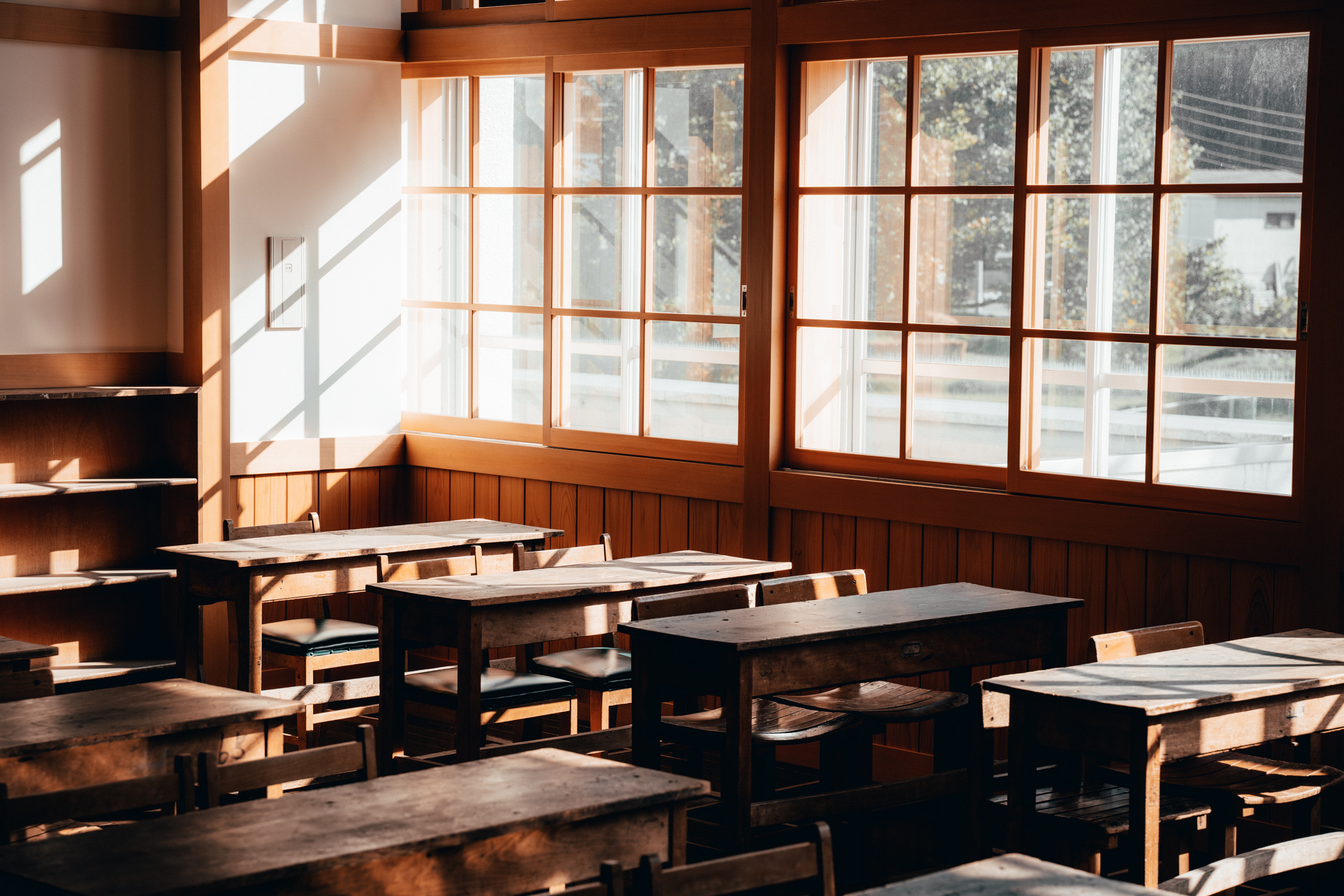 A classroom with old wooden desks and chairs, sun shining in through the windows.