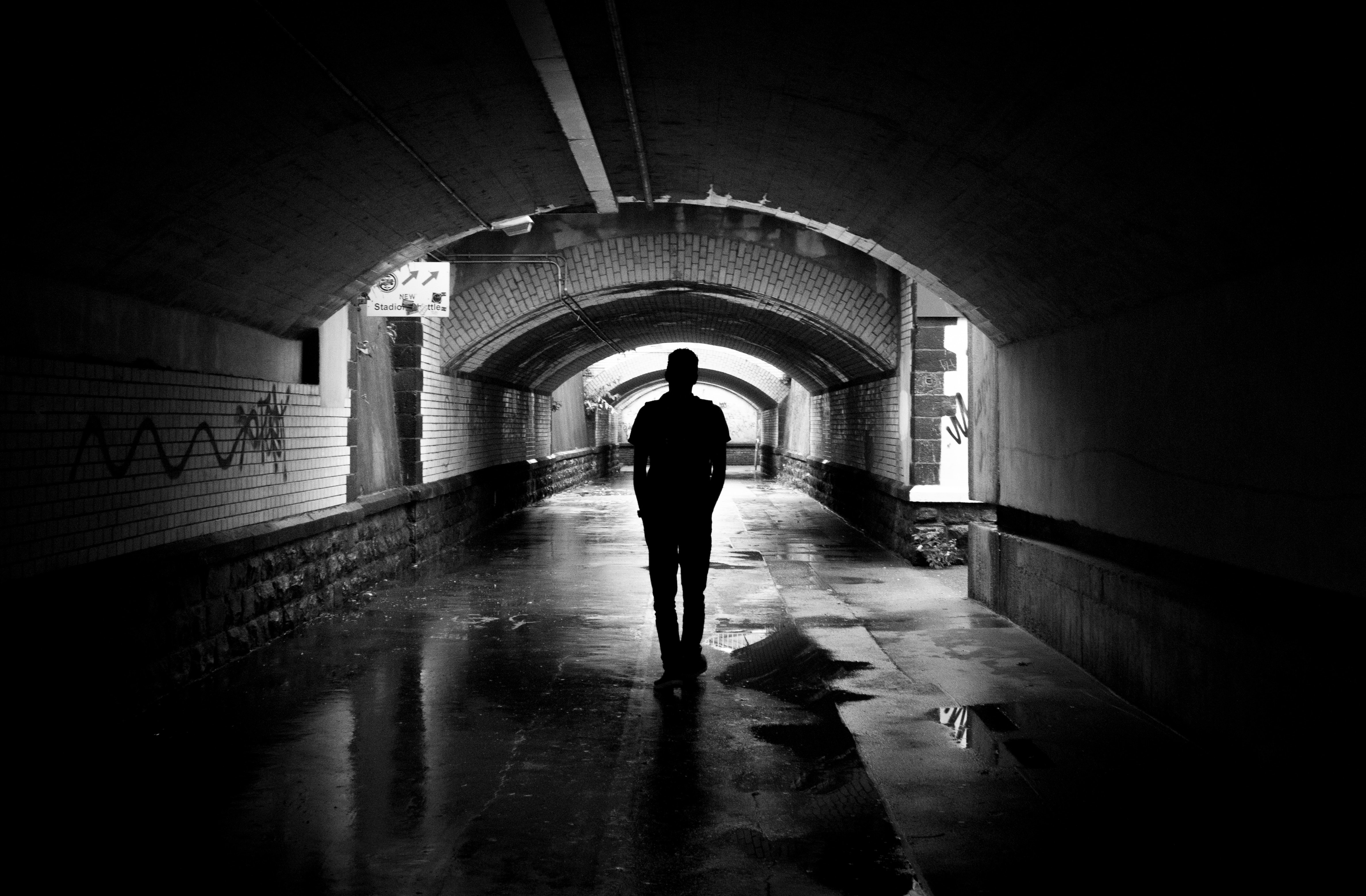 Black and white photo of man walking away from camera through tunnel with graffitied walls. Picture by Lalesh Aldarwish from Pexels.com